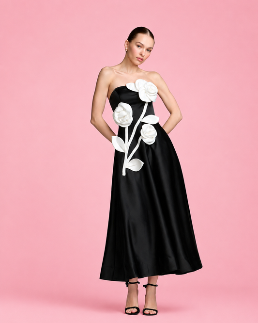 A woman modeling a strapless black satin midi dress with large white 3D rose appliqués on the bodice and skirt, set against a solid pink studio background. Strapless black evening dress with large white floral appliqués