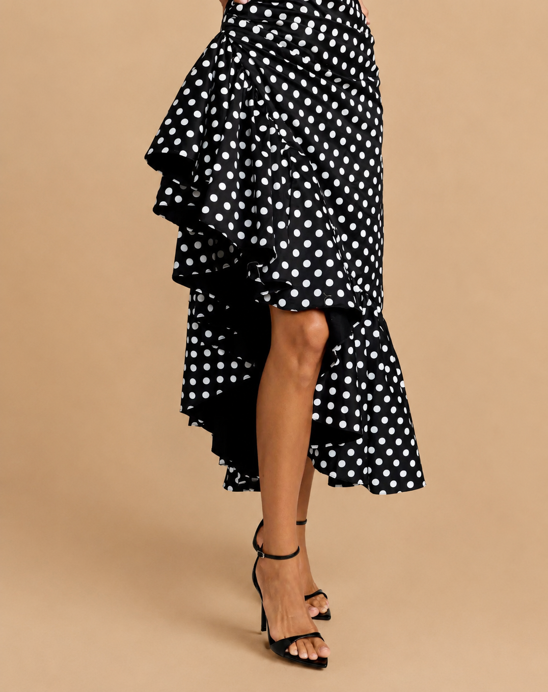 A black and white polka dot ruffled asymmetrical skirt worn by a model, paired with black strappy high-heeled sandals, set against a solid beige studio background.