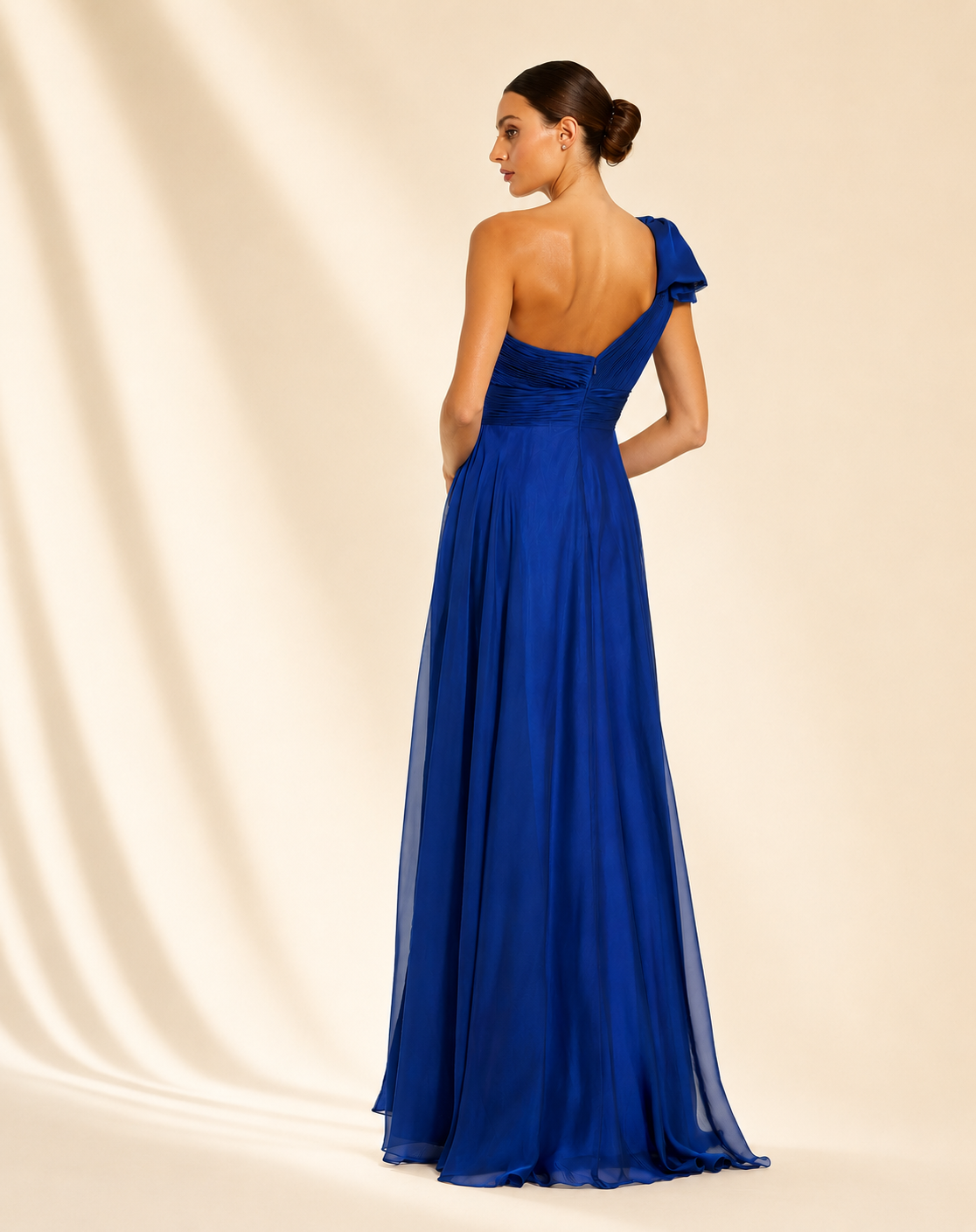 A woman models a floor-length one-shoulder evening gown in a rich royal blue, styled with clear high-heeled sandals against a warm cream studio backdrop. Royal blue one-shoulder floor-length evening gown for women