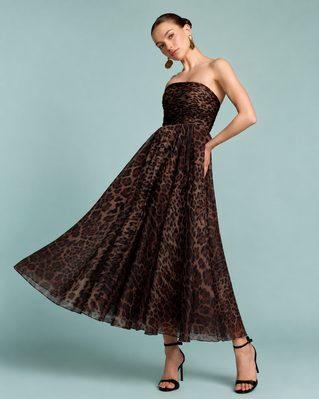 A woman modeling a strapless maxi dress with a brown and black leopard print pattern against a light blue background. Strapless brown and black leopard print maxi dress