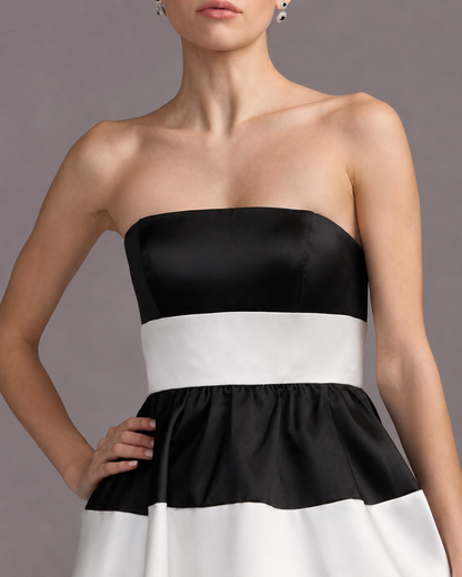 A woman wearing a strapless black and white color-blocked dress made of shiny satin-like fabric, shown from the mouth down against a plain gray studio background. Black and white striped strapless maxi dress for women
