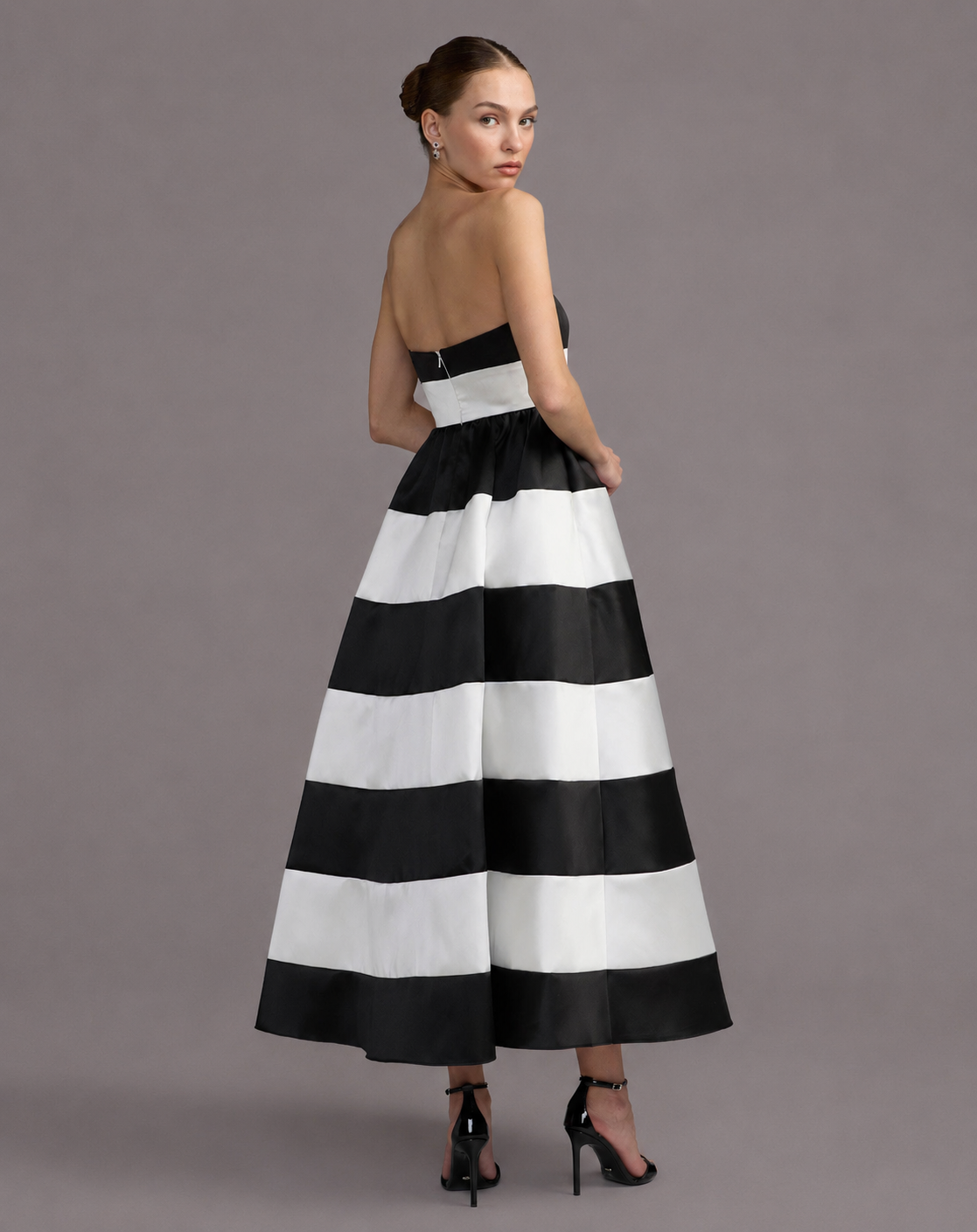 A woman modeling a strapless A-line maxi dress with bold horizontal black and white stripes, shown from the back against a solid gray studio background. Black and white striped strapless maxi dress for women