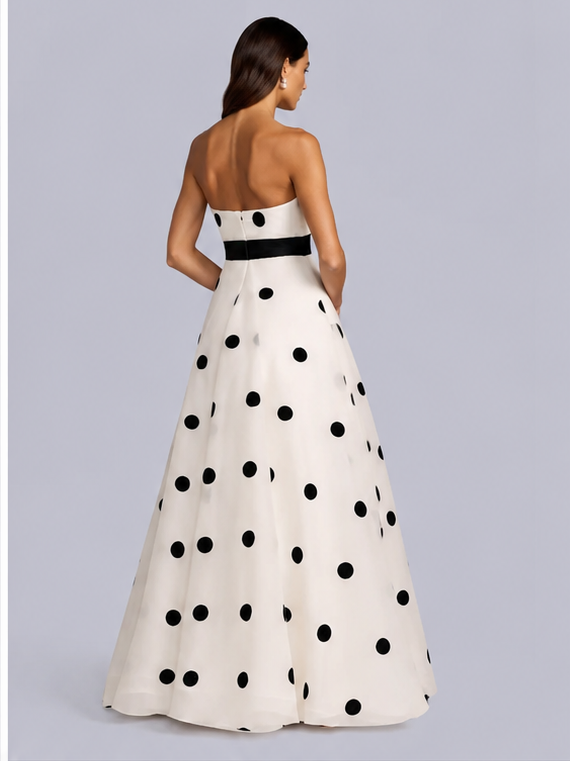 A woman modeling a strapless A-line gown from the back, featuring a white base with large black polka dots and a contrasting black waistband, set against a solid gray studio background.trapless black and white polka dot A-line gown with waist bow