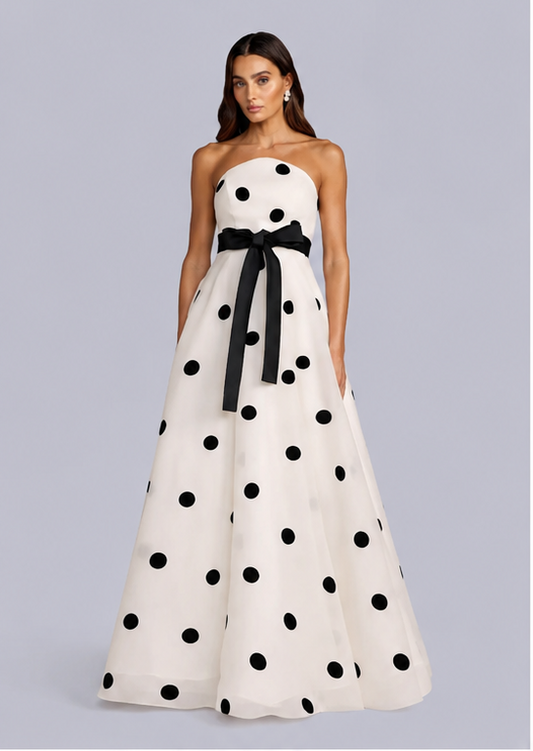 A strapless A-line gown with a black and white polka dot pattern, featuring a wide black waist sash with a bow, modeled against a solid gray background. trapless black and white polka dot A-line gown with waist bow
