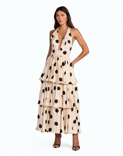 A woman modeling a sleeveless, tiered maxi dress with a beige base and large black polka dots, set against a plain white studio background. Black and beige one-shoulder polka dot maxi dress with bow