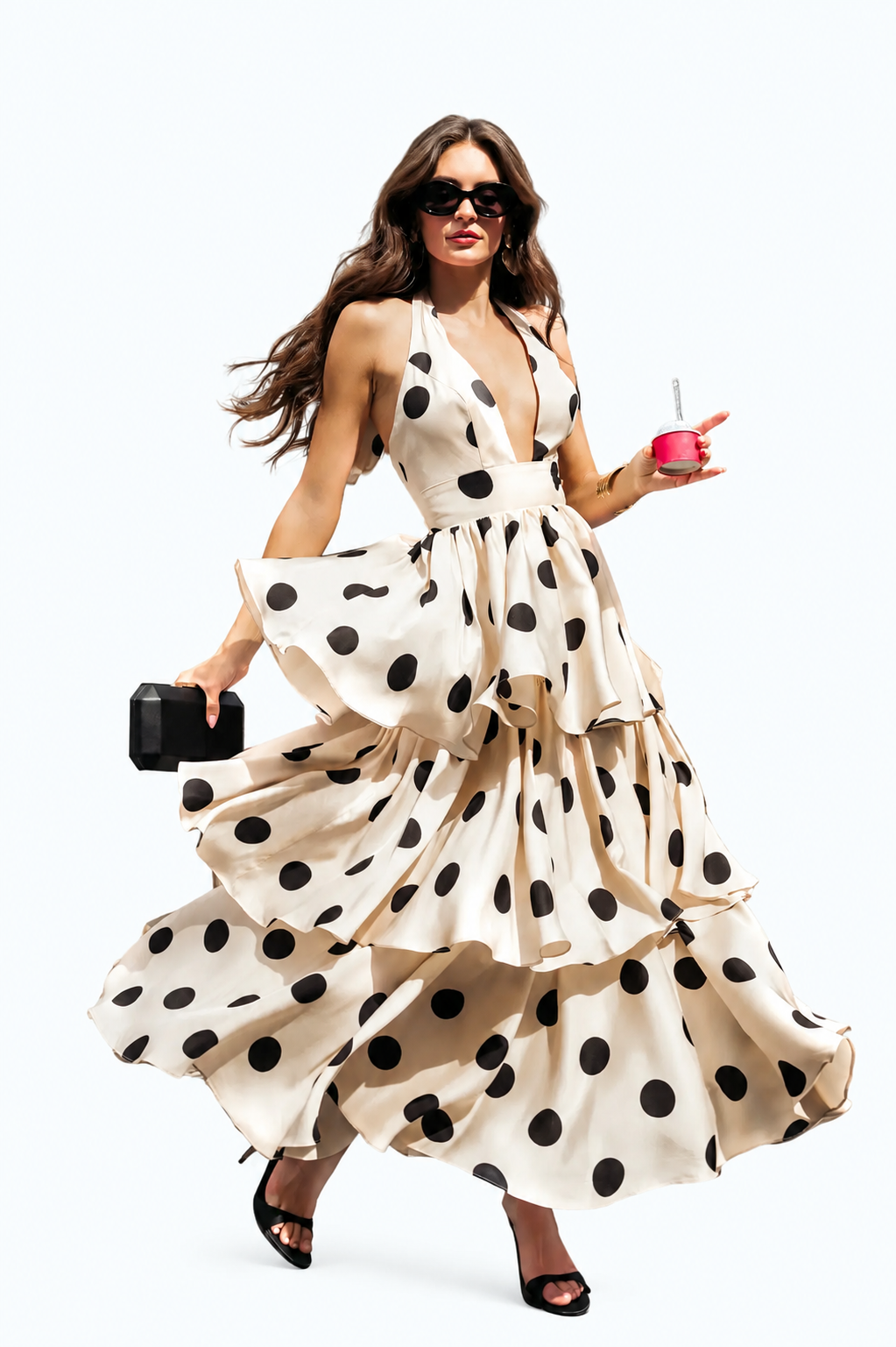 A woman wearing a beige halter-neck maxi dress with large black polka dots, captured mid-motion as she twirls. She is holding a small black clutch and a pink drink, set against a plain white studio background. Black and beige one-shoulder polka dot maxi dress with bow
