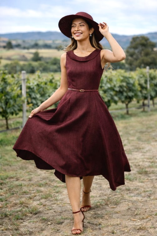 Woman in a burgundy dress and hat standing in a vineyard. Woman wearing burgundy occasion dress in vineyard