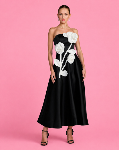 A woman modeling a strapless black satin maxi dress with large white 3D flower appliqués on the bodice and skirt, set against a solid pink studio background. Strapless black evening dress with large white floral appliqués