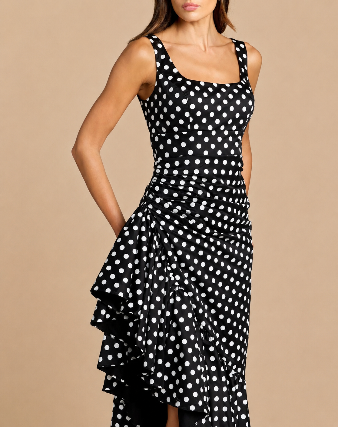 A woman wearing a sleeveless black dress with a white polka dot pattern and a ruffled asymmetrical hem, posed against a solid beige studio background.Black and white polka dot midi dress with asymmetrical ruffle hem
