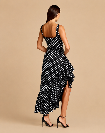 A woman modeling a sleeveless black midi dress with white polka dots, shown from the back against a solid beige studio background. Black and white polka dot midi dress with asymmetrical ruffle hem
