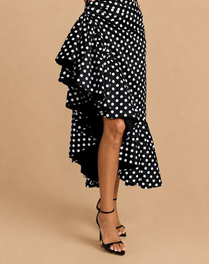 A black and white polka dot ruffled asymmetrical skirt worn by a model, paired with black strappy high-heeled sandals, set against a solid beige studio background.