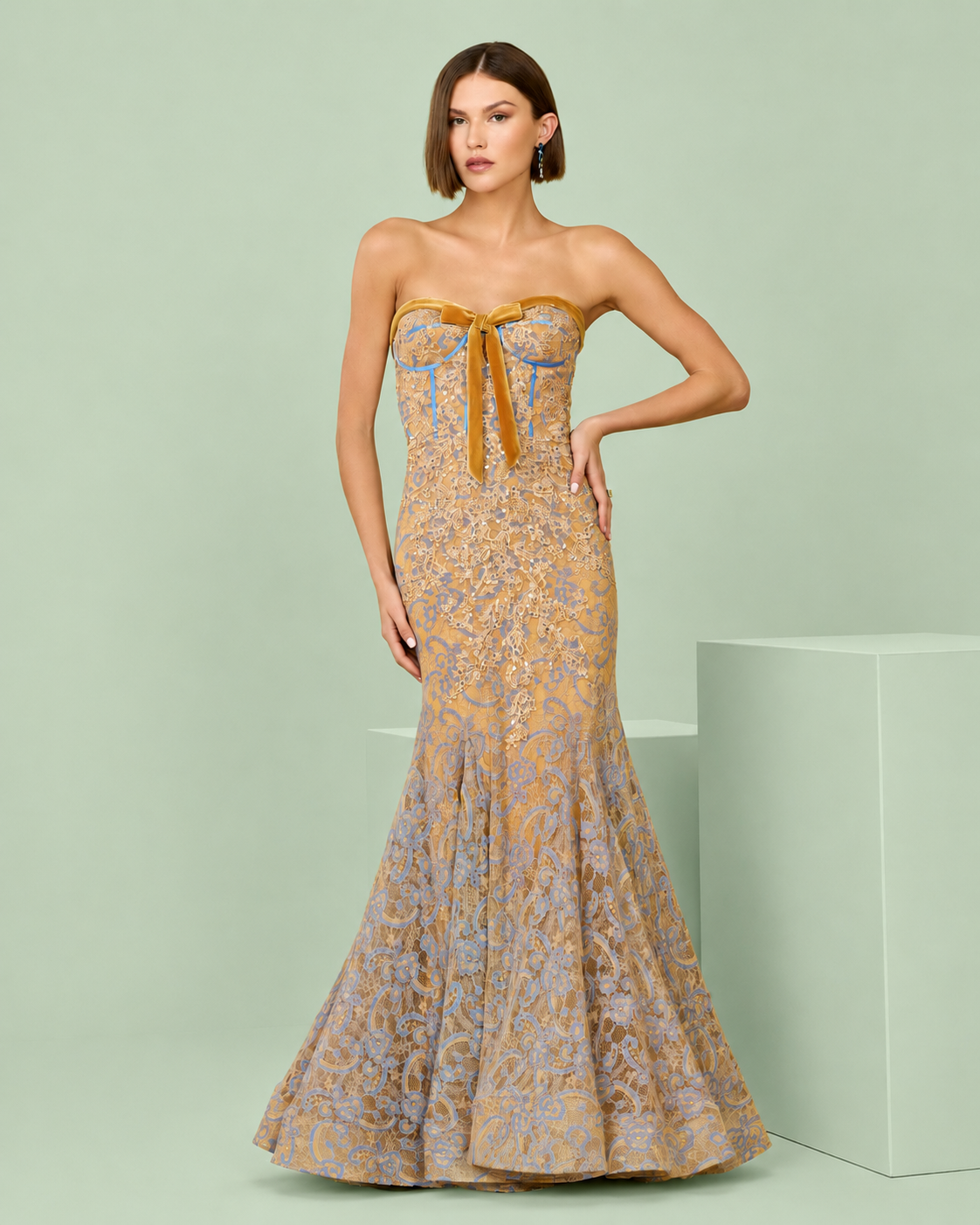 A woman models a strapless mermaid gown with a gold and lavender floral pattern, featuring a satin bow at the sweetheart neckline, set against a light sage green studio background. Gold and lavender floral strapless mermaid gown with satin bow